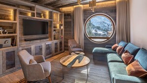 A modern living room with a large flat-screen TV, wooden shelving, a round window with a snowy mountain view, and a cozy seating area with a sofa and armchairs.