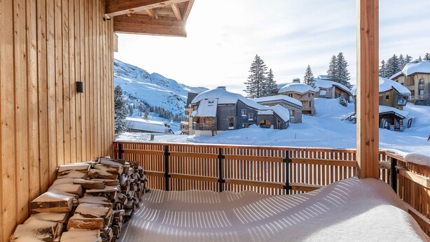 A wooden balcony with a stone wall, overlooking a snowy mountain village with wooden buildings.