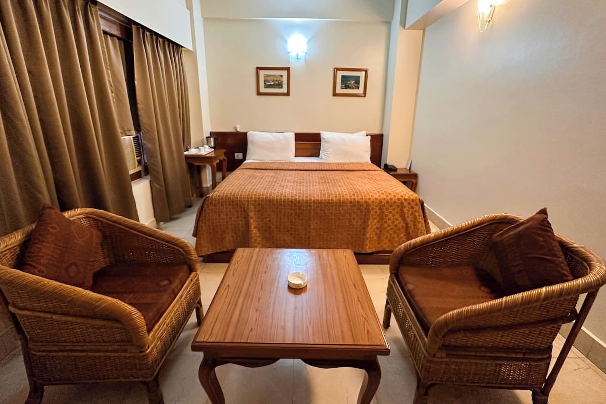 Une chambre d’hôtel avec un lit, deux fauteuils en osier, une table en bois, une table de chevet, une lampe et deux tableaux encadrés accrochés au mur.