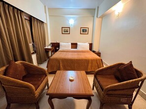 A hotel room with a bed, two wicker chairs, a wooden table, a nightstand, a lamp, and two framed pictures on the wall. - lhaki Hotel (Phuntsholing)