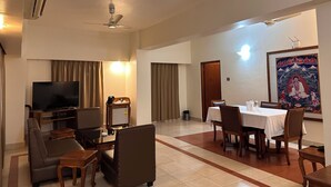 A hotel room with a flat-screen TV, a ceiling fan, a dining area with a table and chairs, and a large framed painting on the wall. - lhaki Hotel (Phuntsholing)