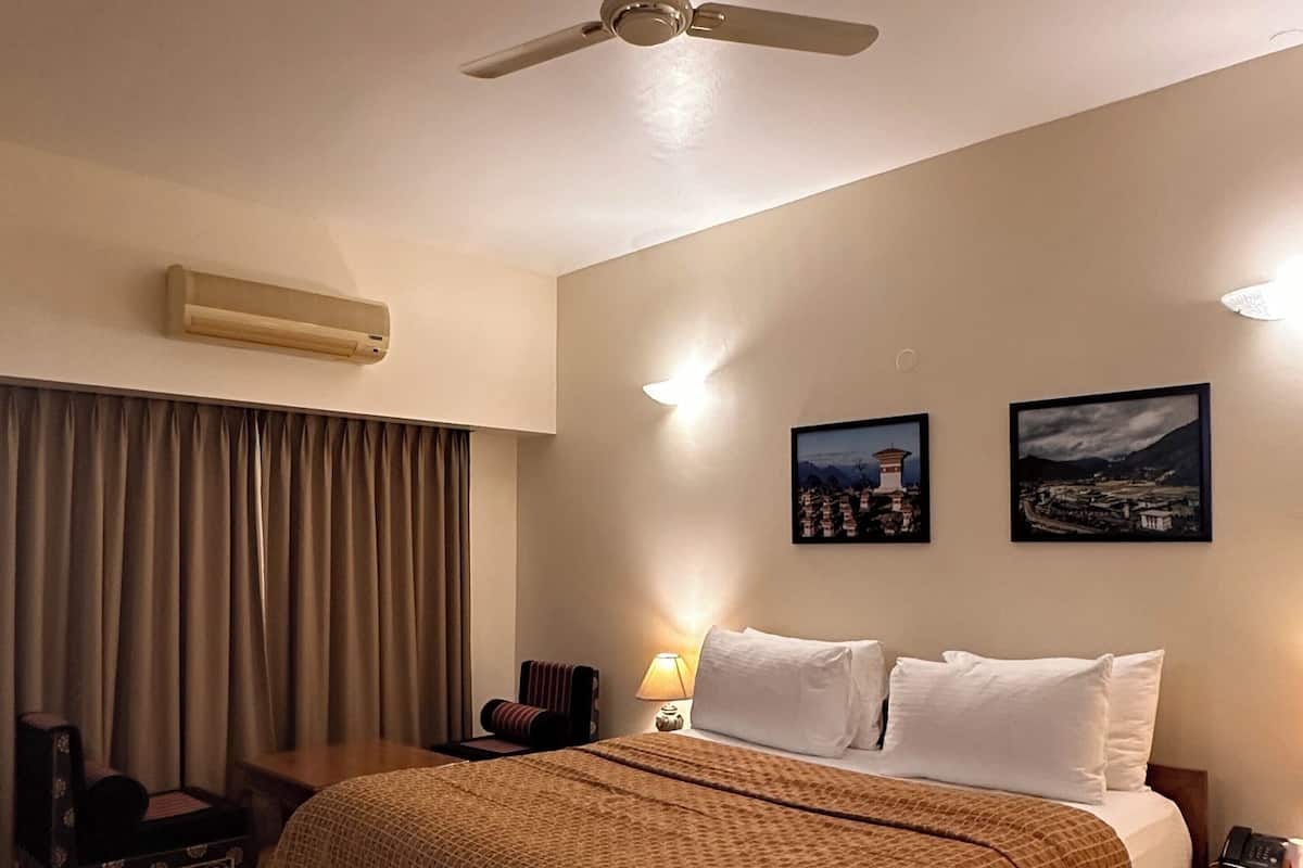 Une chambre d’hôtel comprenant un lit, un ventilateur de plafond, un appareil de climatisation, une table de chevet avec une lampe, une chaise et deux tableaux encadrés accrochés au mur.