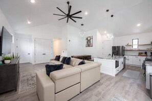 Una sala de estar moderna con un sofá seccional color beige, un ventilador de techo oscuro y una cocina con armarios blancos y encimera de mármol.