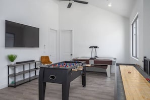 A modern living room with a foosball table, a pool table, a flat-screen TV mounted on the wall, and a ceiling fan.