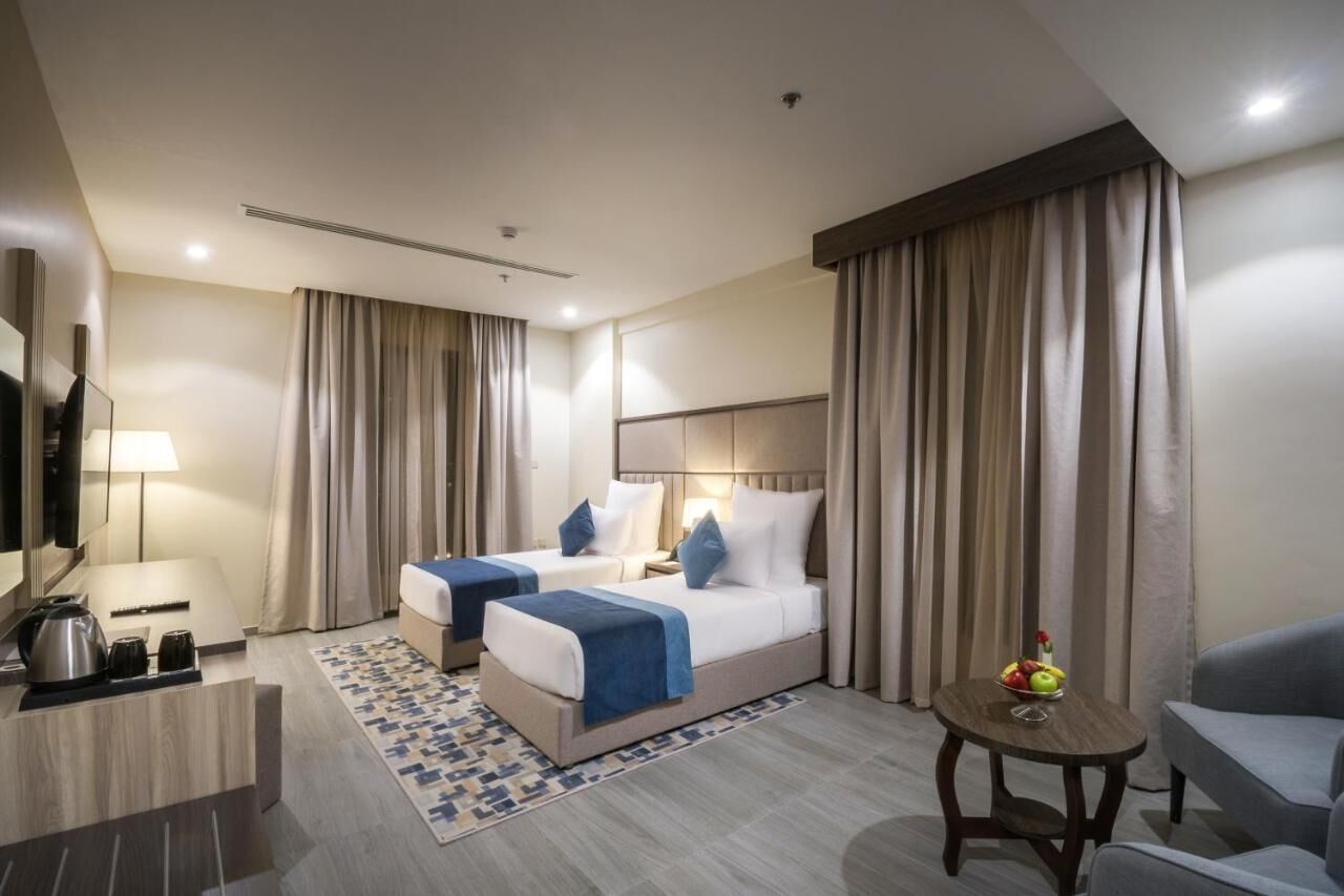 A hotel room with two beds, a desk with a kettle, a small table with fruit, and a TV.