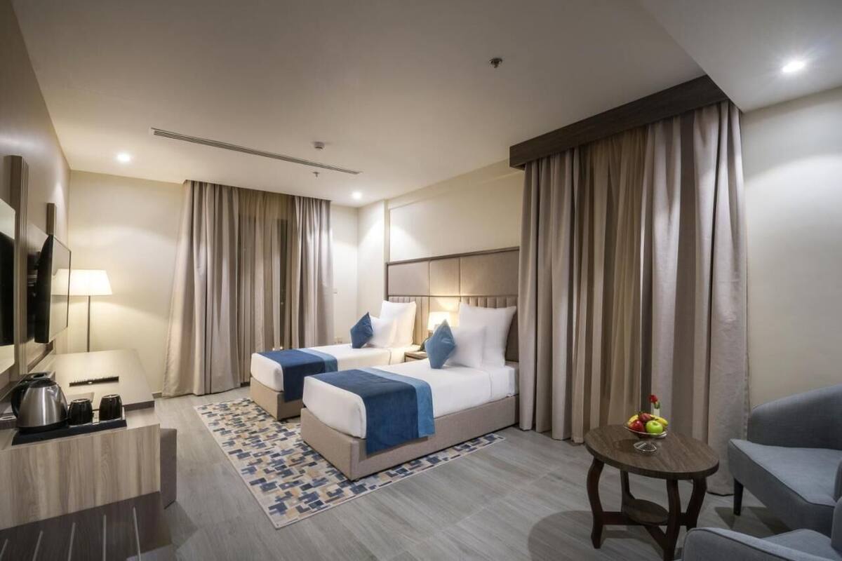 A hotel room with two beds, a desk with a kettle, a small table with fruit, and a TV.