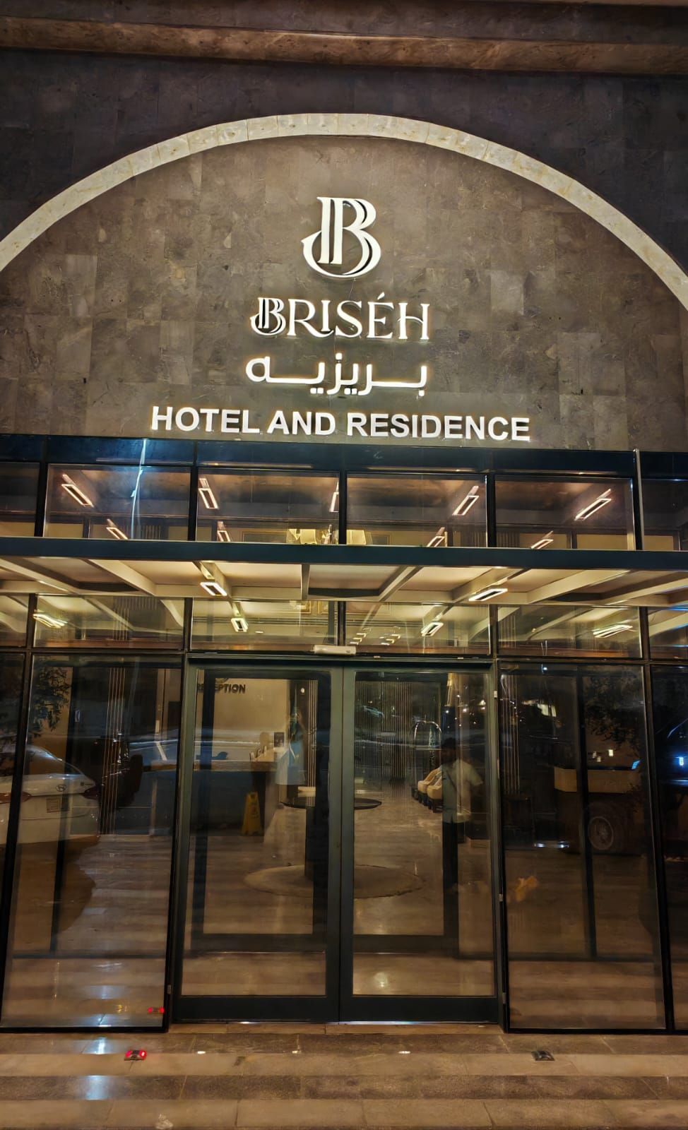 Entrada de un hotel con un amplio arco, puertas de vidrio y un letrero con el nombre del hotel en varios idiomas.