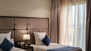 A hotel room with two beds, a nightstand, a lamp, and a view of the city through sheer curtains. - Enala AlKhobar Hotel (Al Khobar)