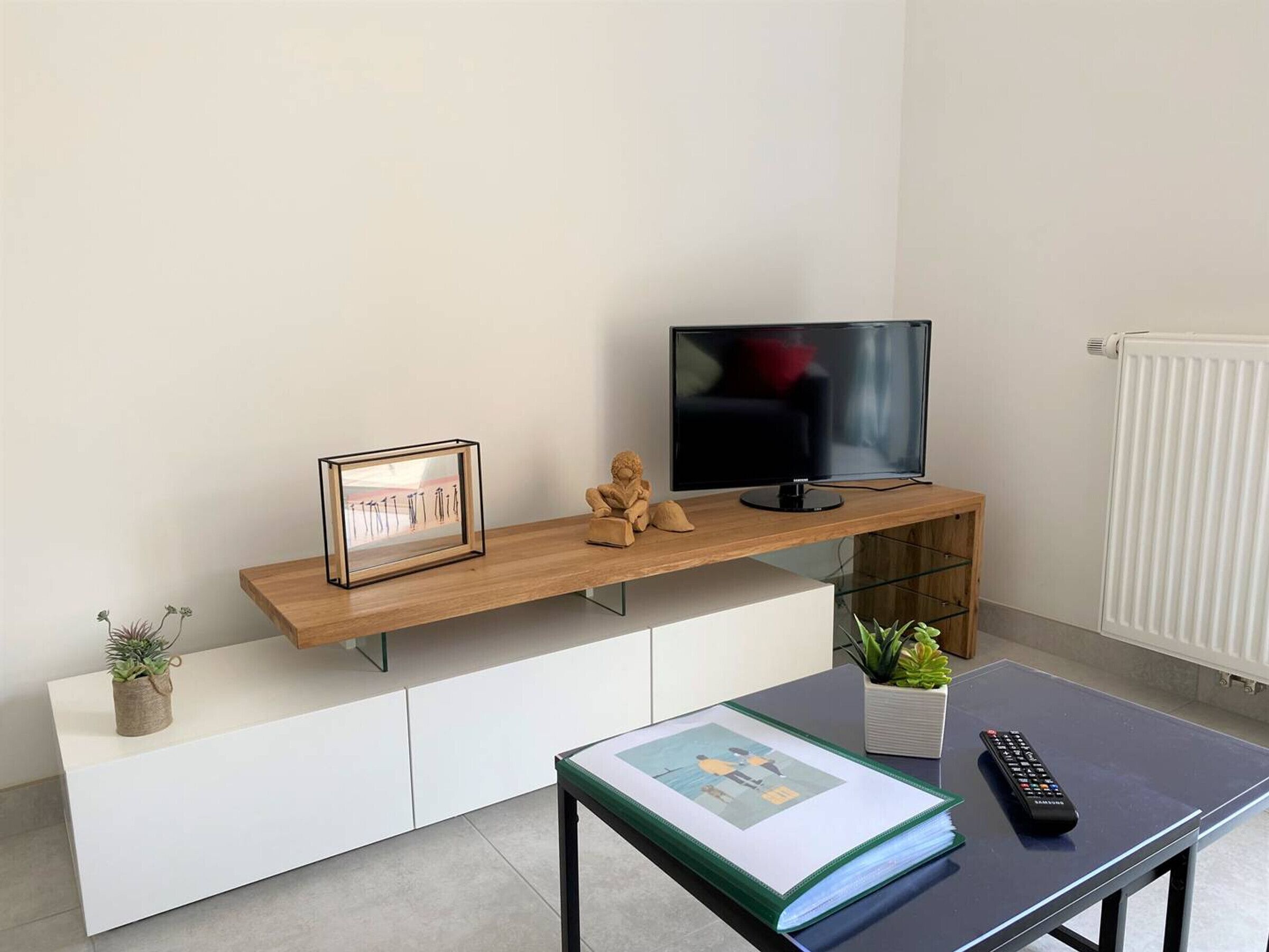 A modern living room with a wooden TV stand, a framed picture, a small statue, a remote control, and a book on a table.