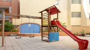 A playground with swings and a slide, surrounded by a tiled patio and a building with a stone wall and windows. - Central Quiet Apartment with Pool, Wi-Fi in Torredembarra (Torredembarra)