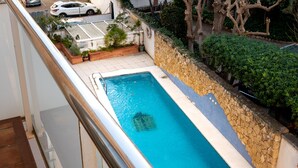 A rectangular swimming pool with a stone wall and tiled deck, surrounded by greenery and a parking area in the background. - Central Quiet Apartment with Pool, Wi-Fi in Torredembarra (Torredembarra)