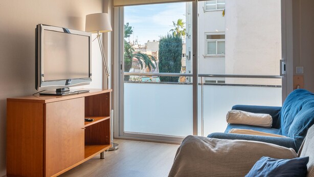 A modern living room with a flat-screen TV on a wooden cabinet, a sofa with cushions, and a large sliding glass door leading to an outdoor balcony. - Central Quiet Apartment with Pool, Wi-Fi in Torredembarra (Torredembarra)