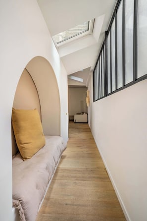 A narrow hallway with a high ceiling, a skylight, a cushioned bench with a yellow pillow, and a white wall with a window.