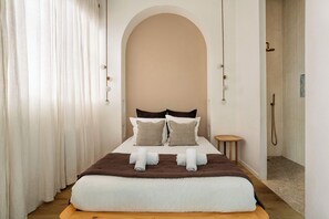 Quarto de hotel com uma cama grande, duas almofadas, cabeceira de madeira, uma mesa redonda pequena e área de chuveiro.
