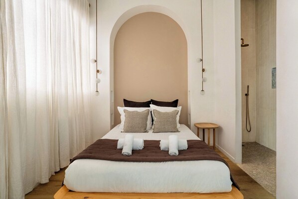 A hotel room with a large bed, two pillows, a wooden headboard, a small round table, and a shower area.