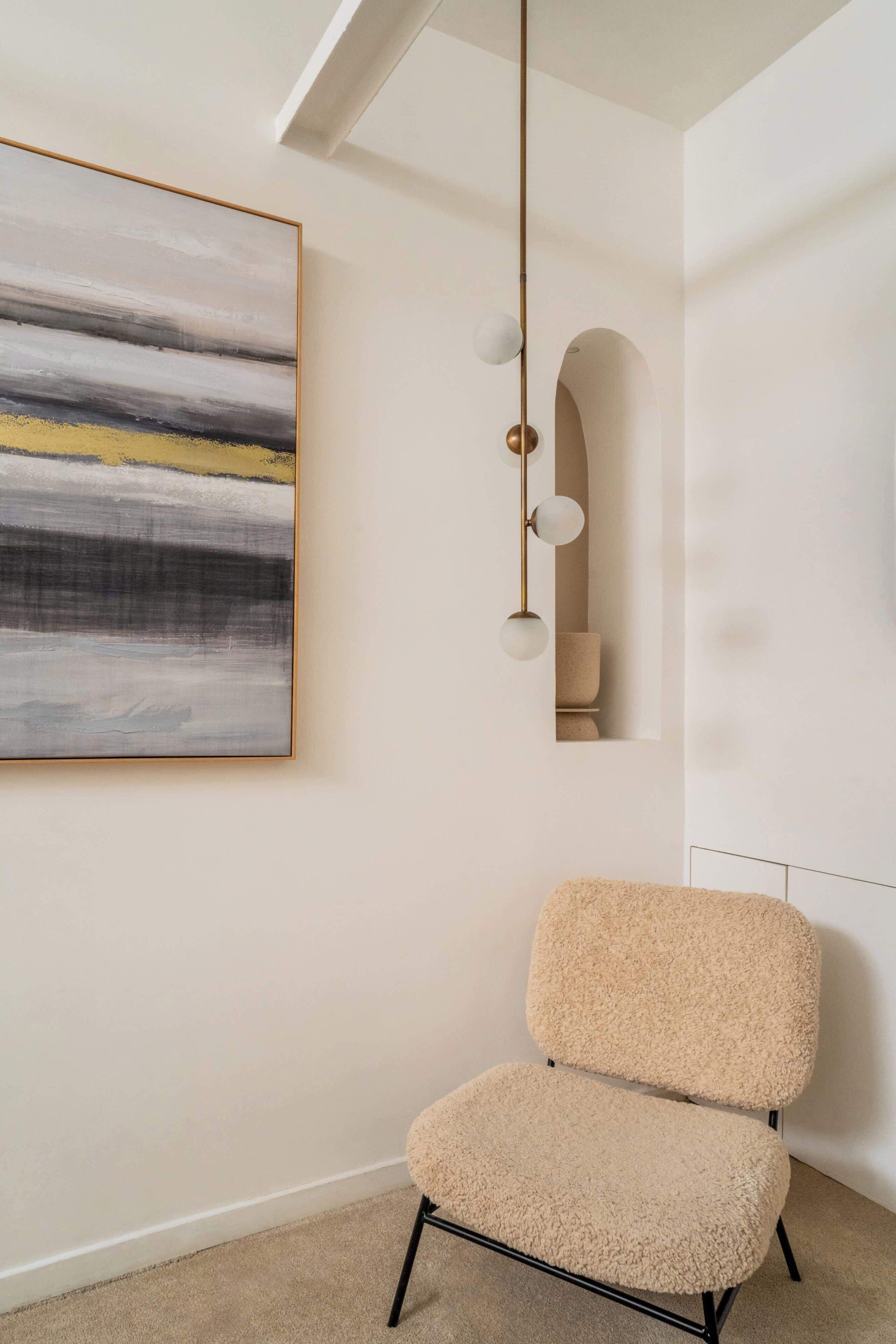 A room with a minimalist design, featuring a beige chair, a modern light fixture, and a large abstract painting on the wall.