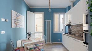 A modern kitchen with white cabinets, a gas stove, and a window. There is a dining table with chairs and a radiator in the background. - PETRONIUS HOME STUDIO (Bologna)