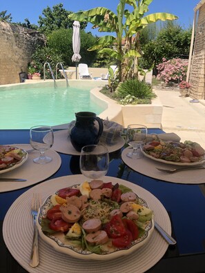 A dining table with plates of food, a pitcher, and glasses, set outdoors with a pool and plants in the background.