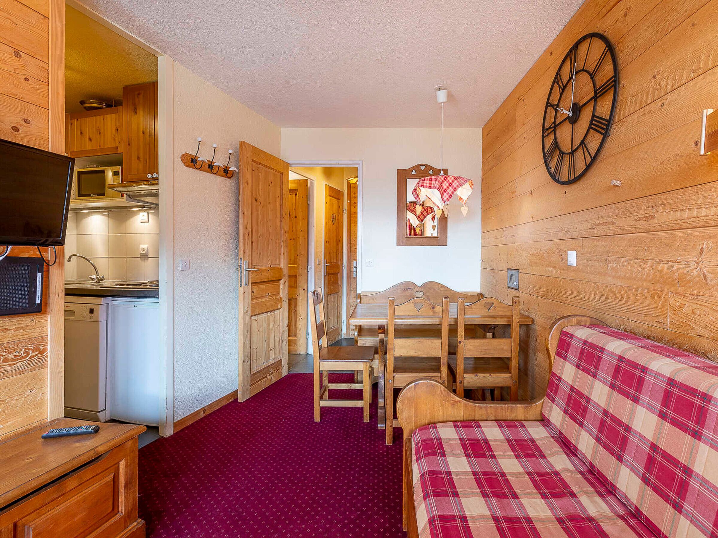 Et kompakt værelse med et træspisebord, en rød ternet sofa, et ur og et tekøkken med en mikroovn.