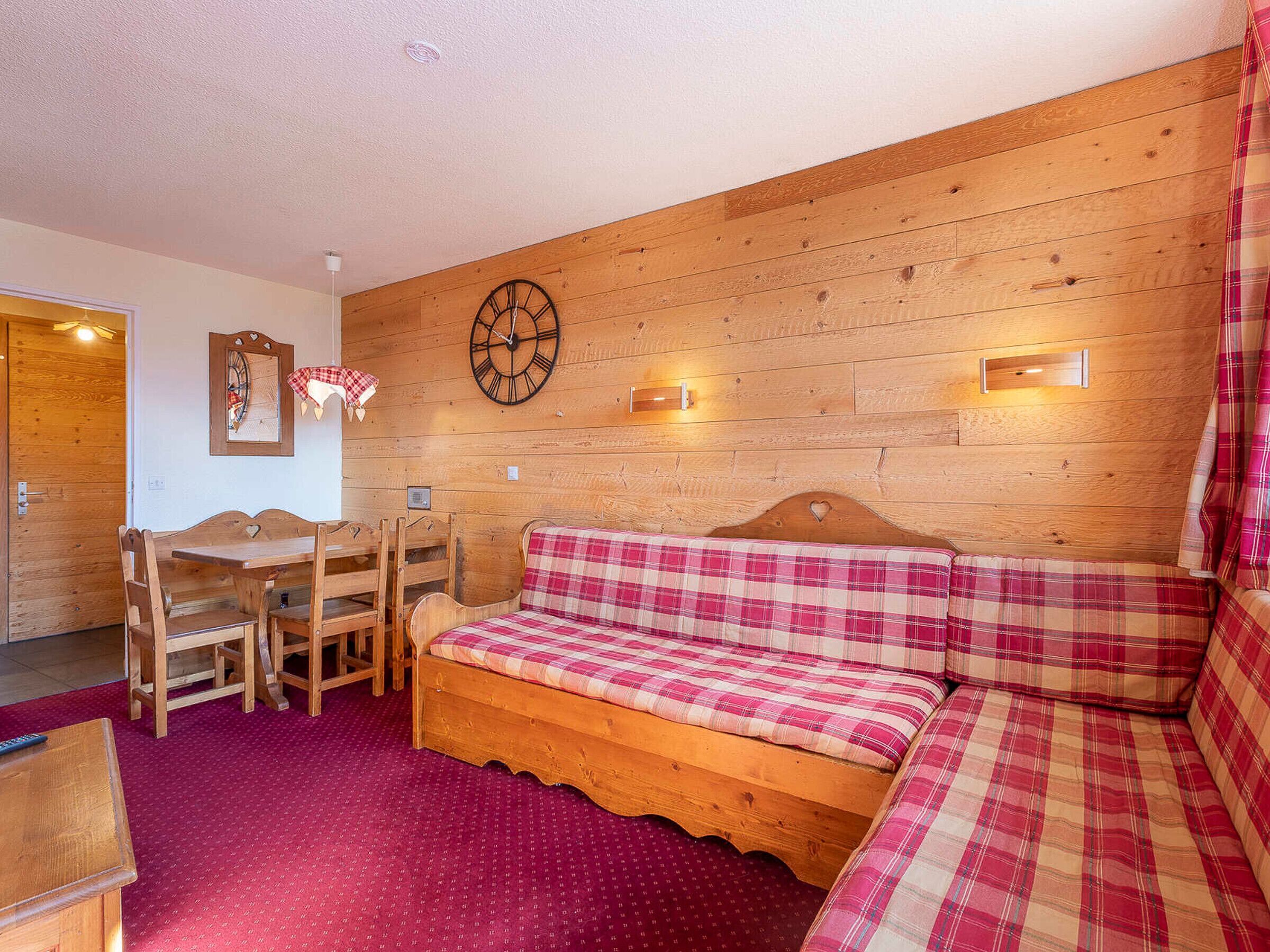 Une pièce chaleureuse avec un mur en bois, une horloge murale ronde, un banc en bois agrémenté d’un coussin à carreaux et une table avec des chaises.