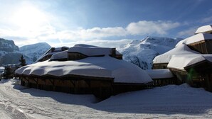 Un bâtiment en bois recouvert de neige, avec un toit également enneigé, entouré de montagnes enneigées et surplombé d’un ciel dégagé.