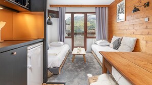 Une chambre d’hôtel compacte dotée d’une kitchenette, de deux lits simples, d’une table en bois et offrant une vue sur les montagnes grâce à de grandes fenêtres.