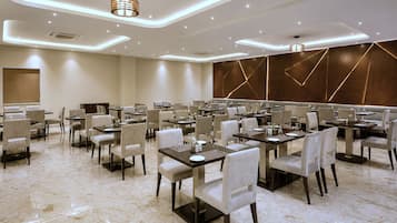 Un comedor de hotel moderno con varias mesas y sillas, iluminación empotrada en el techo y una pantalla grande en la pared.