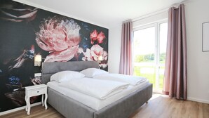 Quarto com cama cinza, roupa de cama branca, uma mesinha de cabeceira pequena e branca, um grande mural floral na parede e vista para o verde pela janela.