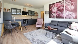 Uma sala de estar moderna com um sofá cinzento, uma mesa de centro e uma poltrona rosa. Há também uma área de cozinha com armários e uma televisão.