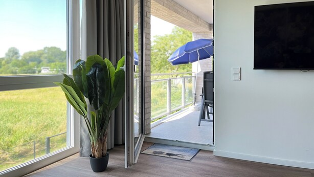 Quarto com uma grande janela, uma planta em vaso e vista para uma varanda com um guarda-sol azul.