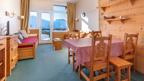 Une pièce confortable avec un lit, un bureau et une table à manger avec des chaises. On y trouve des fenêtres avec des rideaux, une horloge et une étagère avec des objets décoratifs.