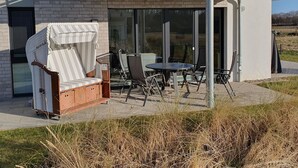 Ein moderner Außenbereich mit Sitzgelegenheiten, darunter ein Korbstuhl, ein Tisch und Stühle, neben einem Gebäude mit großen Fenstern und einer Glastür.