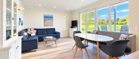 Un salon moderne avec un canapé bleu, une table à manger avec des chaises et un téléviseur. Une grande fenêtre offre une vue sur l’extérieur.