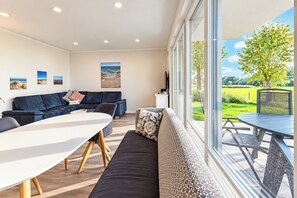 A modern living room with a sofa, a dining table, and a large window offering a view of a garden.