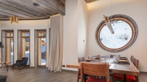 Uma sala de jantar com uma mesa redonda, cadeiras de madeira e vista para uma paisagem nevada através de uma grande janela.