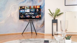 A modern living room with a flat-screen TV on a tripod, a dining table with a remote control, a potted plant, and a radiator. - Penthouse-Suite near University (Gießen)