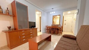 Un salon comprenant un canapé, un meuble TV en bois, une table à manger avec des chaises, et une cuisine visible à travers une porte ouverte.