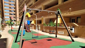 Un terrain de jeux équipé de balançoires et de toboggans, entouré d’un immeuble à plusieurs étages doté de balcons.