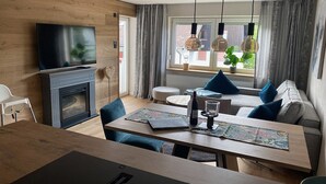 A modern living room with a fireplace, a dining area with a table and chairs, a sofa with cushions, and a TV mounted on the wall. - Willkommen in der Gemütlichen Ferienwohnung Bergidyll (Oberstaufen)