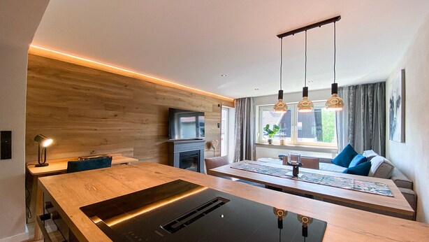 A modern kitchen with a central island, wooden wall paneling, a built-in TV, and pendant lighting. - Willkommen in der Gemütlichen Ferienwohnung Bergidyll (Oberstaufen)