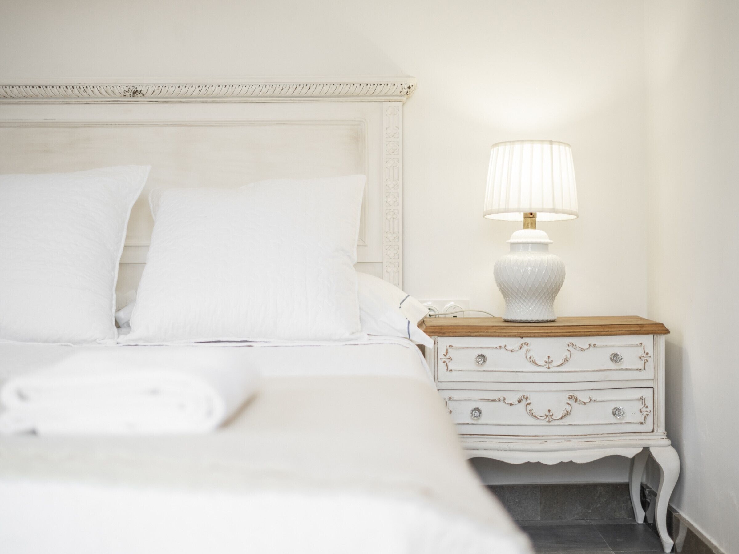 A neatly made bed with white linens, a bedside lamp, and a white dresser with drawers.