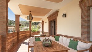 Un patio techado con una mesa de madera y un sofá, paredes de ladrillo y vistas a las colinas.
