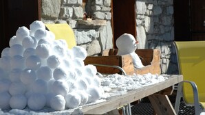 A pile of snowballs and a snowman figure on a wooden table outdoors, with stone walls and a yellow chair in the background. - Comfortable 3-star apartment, large terrace and private parking (Le Corbier)