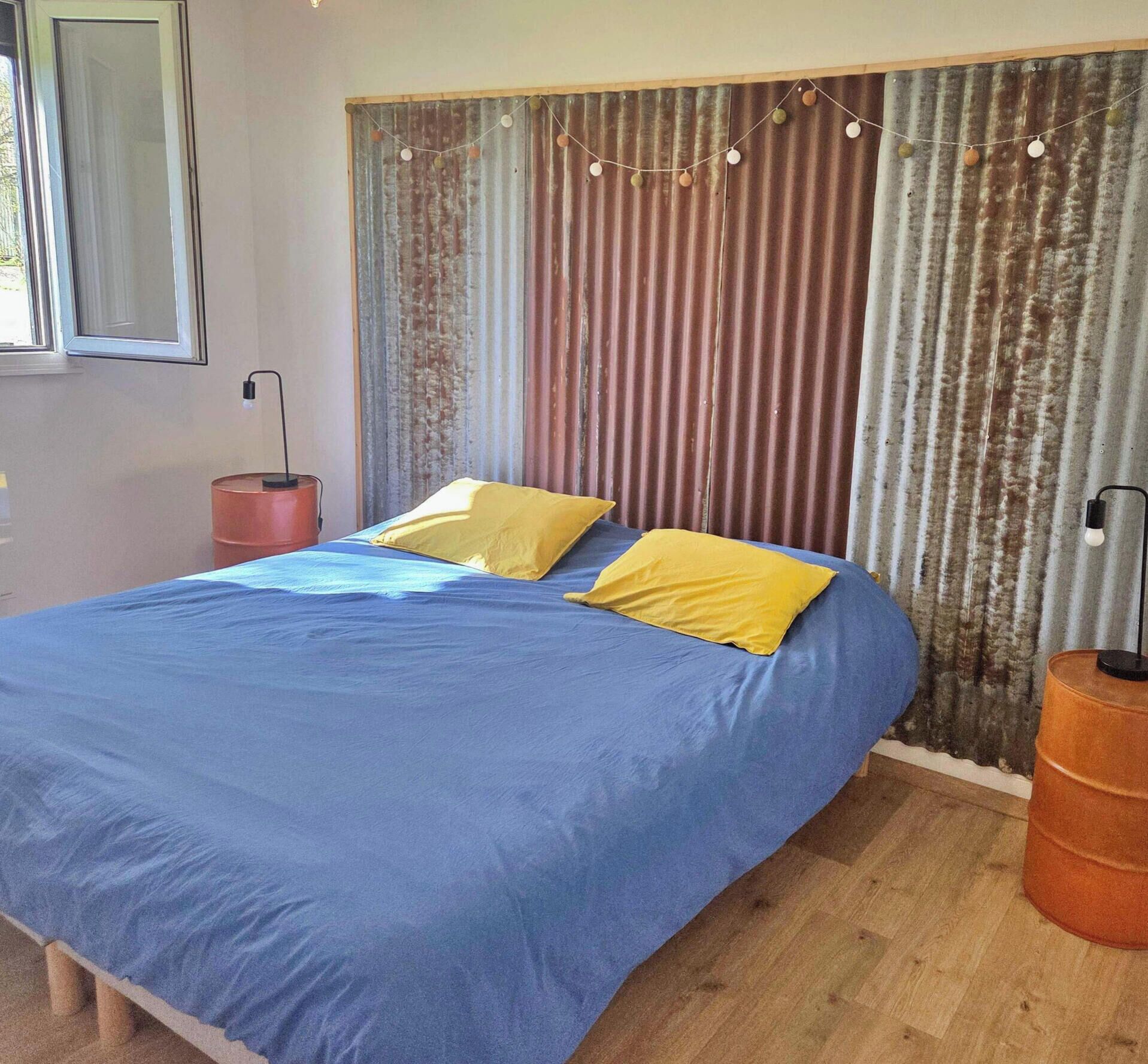 Ein Schlafzimmer mit einem großen Bett, zwei Kissen, einer Lampe, einem Mülleimer und einem Fenster.