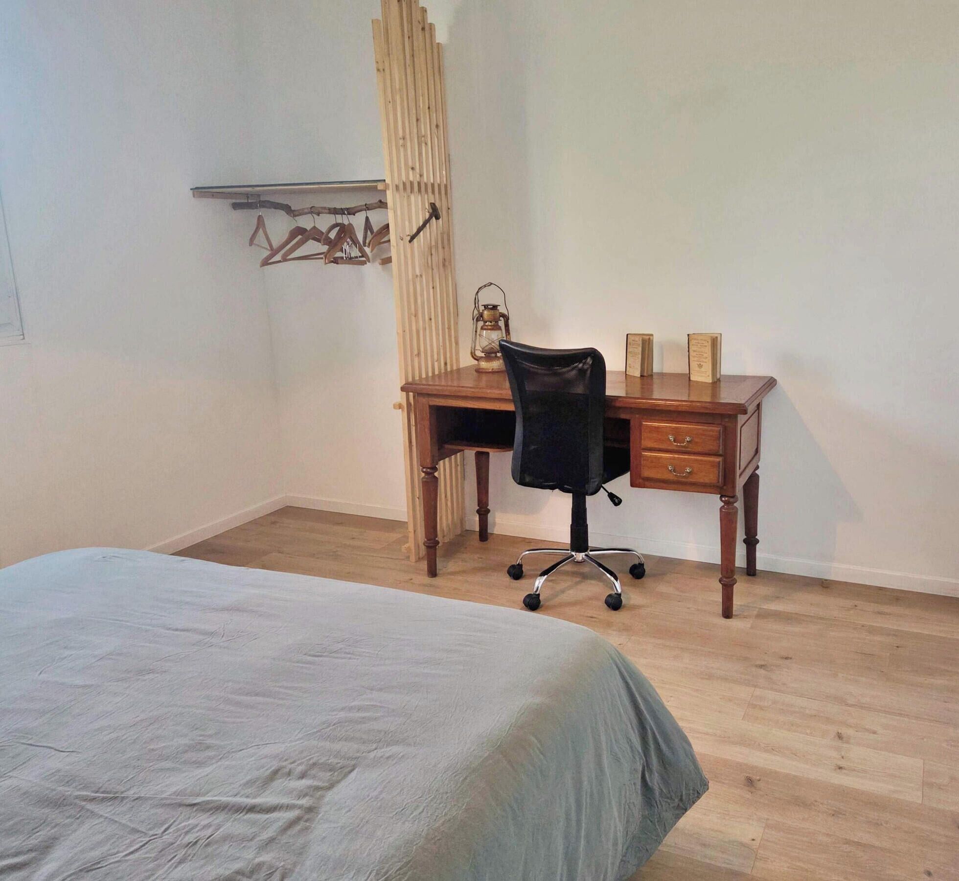 A bedroom with a bed, a wooden desk, a black office chair, and a wooden wardrobe.