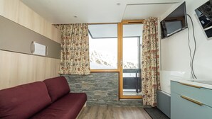A room with a wooden floor, a red sofa, a small kitchen area with a TV, and a window with curtains. - Comfortable studio ski-in/out with balcony and WIFI (Tignes)