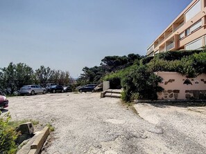 A parking lot with cars, a building with balconies, and a stone wall with vegetation.