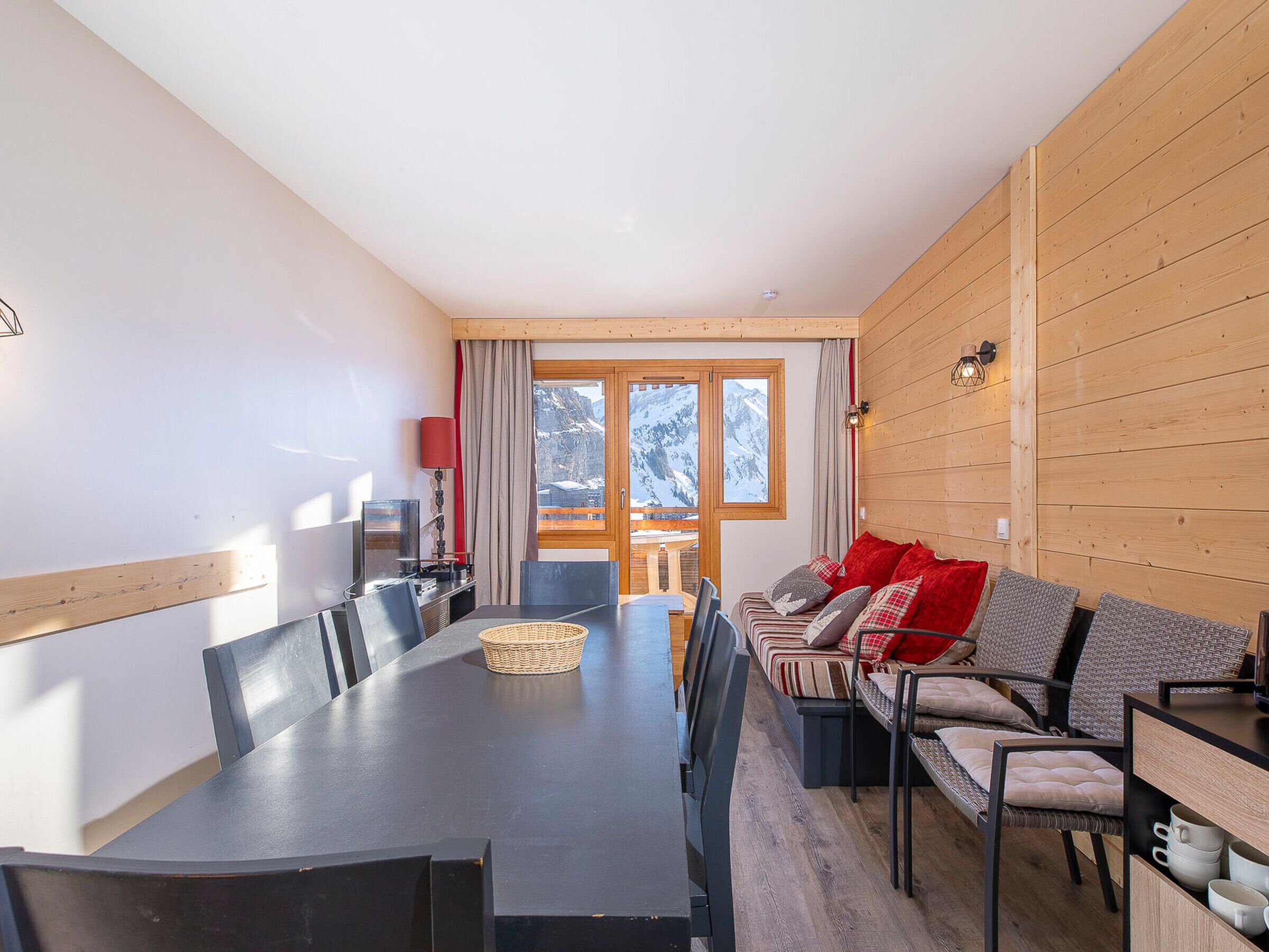 A modern dining area with a long table, multiple chairs, and a sofa with red cushions.