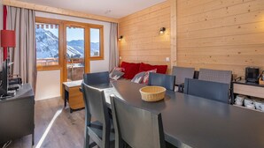 A modern room with a wooden interior, a dining table with chairs, a sofa with red cushions, and a view of snow-capped mountains through large windows.