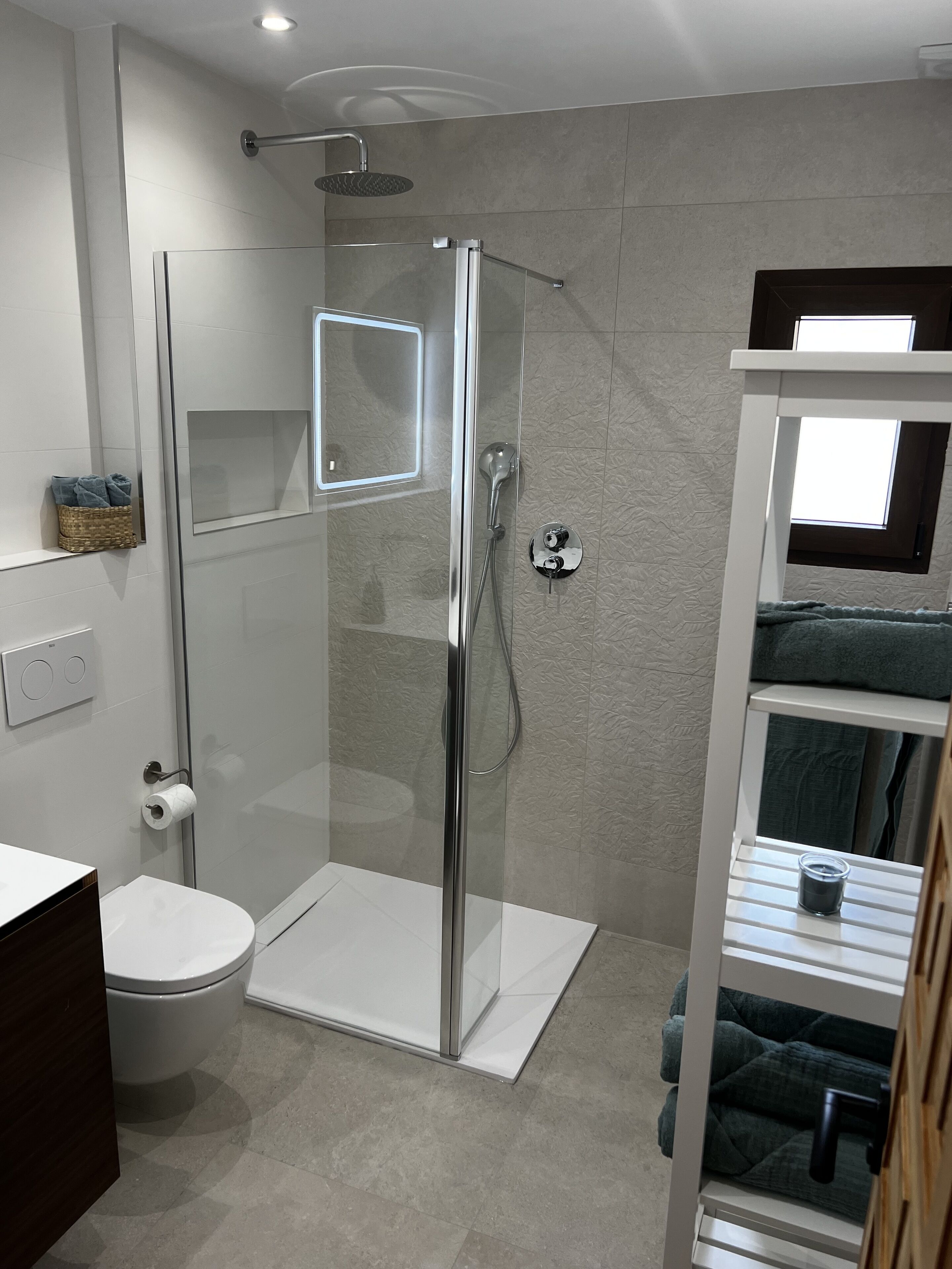 A modern bathroom with a glass-enclosed shower, a toilet, and a built-in shelf with towels.